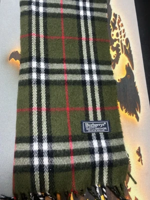 Grön rutig halsduk från Burberrys - Snygg halsduk i klassiskt rutigt mönster från Burberrys. Färgen är olivgrön med svarta, vita och röda ränder. Halsduken är tillverkad i 100% lammull och har fransar i ändarna. Perfekt accessoar för att lyfta din outfit.