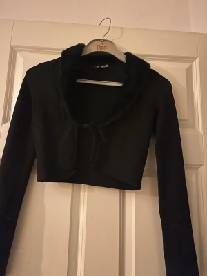 Svart kort kofta från H&M Divided - Snygg svart kort kofta från H&M Divided med långa ärmar och mjuk fuskpäls runt kragen. Koftan knyts framtill och har en tight passform som passar perfekt till lager på lager-stil. Materialet är stretchigt och bekvämt.
