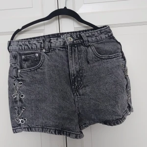 Grå jeansshorts med kedjedetaljer FB Sister - Säljer ett par grå, tvättade jeansshorts från FB Sister i storlek M. Shortsen har hög midja och coola kedjedetaljer på sidorna som ger en edgy vibe. Klassisk femficksmodell och knappstängning framtill. Perfekta för dig som vill sticka ut lite extra.