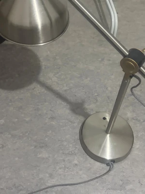 Bordslampa i borstat stål - Snygg bordslampa i borstat stål med rund fot och justerbar arm. Lampan har en klassisk, industriell design och en textilklädd sladd i svartvitt mönster. Perfekt för skrivbord eller nattduksbord.