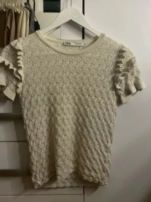 Beige topp med volangärm från Zara - Supersöt beige topp från Zara med korta volangärmar och ett stickat, strukturerat mönster över hela plagget. Rund halsringning och mjukt material som känns skönt mot huden. Perfekt för dig som gillar en romantisk och trendig stil.
