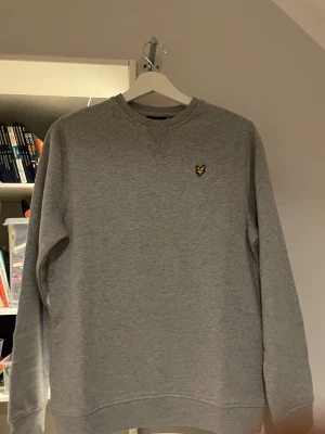 Grå sweatshirt från Lyle & Scott - Klassisk grå sweatshirt från Lyle & Scott med rund halsringning och den ikoniska gula örnloggan broderad på bröstet. Tröjan har ribbade muddar vid ärmslut och nederkant. Perfekt basic-plagg i mjukt bomullsmaterial. 158-164