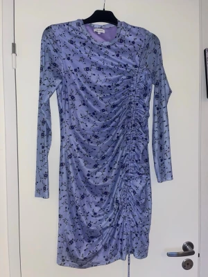 Ljuslila blommig klänning - Supersnygg ljuslila kortklänning från Bubbleroom med blommigt mönster och rynkade detaljer längs sidan. Klänningen har lång ärm, rund halsringning och snörning i sidan för extra cool look. Perfekt för dig som vill sticka ut lite extra!