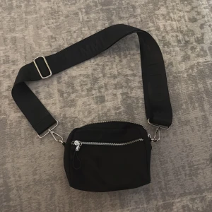 Svart väska  - Snygg svart magväska  med bred justerbar axelrem där det står 'FEMME FORTE'. Väskan har två fack med silverfärgade dragkedjor och är tillverkad i ett slitstarkt tygmaterial. Perfekt för att bära det viktigaste på ett stilrent sätt.