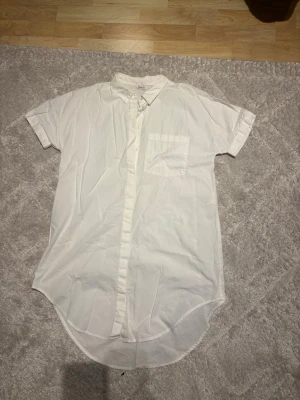 Vit kortärmad skjorta från ONLY - Klassisk vit skjorta från ONLY med korta ärmar och bröstficka. Skjortan har knappar framtill och en avslappnad passform. Är väldigt lång i passformen. Perfekt till jeans eller shorts för en clean look.