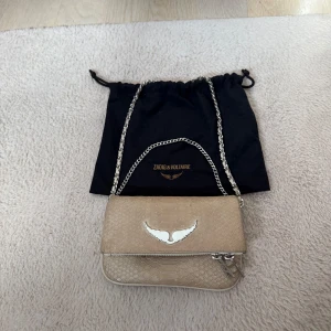 Beige Zadig & Voltaire väska - Snygg beige axelväska från Zadig & Voltaire med kedjerem och silverfärgat emblem framtill. Väskan har ett reptilmönstrat skinn och dragkedja både fram och bak. 