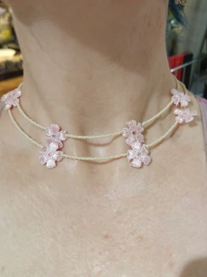 Choker med pärlor och rosa blommor - Dubbel choker med små vita pärlor och broderade ljusrosa blommor. Halsbandet har två rader och blommorna är jämnt placerade längs hela halsbandet. Perfekt för dig som gillar söta och romantiska accessoarer.