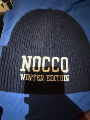 Mörkblå ribbstickad mössa NOCCO - Säljer en mörkblå ribbstickad mössa från NOCCO med texten 'WINTER EDITION' broderad framtill. Klassisk uppvikt kant och mjukt material som håller dig varm under kalla dagar. Perfekt accessoar för vintern och snygg till streetwear.