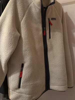 Vit/begie patagonia fleece - Mysig fleece från Patagonia med dragkedja framtill och svarta detaljer. Jackan har tre fickor med röda dragkedjor och en hög krage. Tillverkad i fluffigt teddy-material som håller dig varm och bekväm. Perfekt för kyliga dagar.