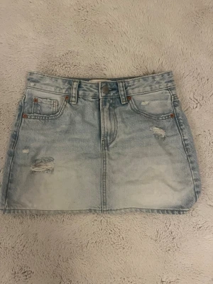 Ljusblå jeanskjol från Stradivarius - Trendig ljusblå jeanskjol från Stradivarius med slitna detaljer och klassiska fem fickor. Kjolen har bälteshällor och knappstängning framtill. Perfekt till sommaren och enkel att matcha med olika toppar.