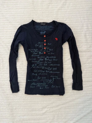Sällsynt Abercrombie & fitch Henley  - Sällsynt vintage Henley-tröja från Abercrombie & Fitch i mörkblått, långärmad med grafiskt tryck.  Storleken är okänd men passar ungefär XS. Mycket gott skick – varsamt använd utan synliga fläckar eller skador.  Mörkblå Henley-tröja från Abercrombie & Fitch med ett unikt script-stil grafiskt tryck över framsidan och klarröda knappar vid halsringningen.  Knapparna går tyvärr inte att öppna eller justera.