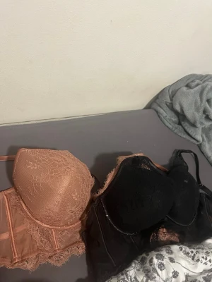 Två spetsbhar i svart och nude - Säljer två snygga bhar med spetsdetaljer, en i svart och en i nude/ljusbrun färg. Båda har vadderade kupor, justerbara axelband och dekorativ snörning framtill. Materialet är en kombination av spets och mesh som ger en lyxig känsla.