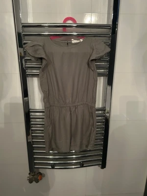 Grön jumpsuit med volangärmar - En stilren olivgrön jumpsuit med volangärmar och rund hals. Jumpsuiten har markerad midja och är tillverkad i ett mjukt material som ger en skön passform. Perfekt för dig som gillar enkel och modern design.