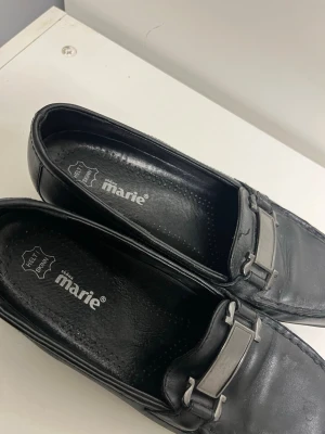 Svarta loafers från Marie i skinn - Stilrena svarta loafers från Marie i äkta skinn med metallspänne över vristen. Klassisk design med fyrkantig tå och diskreta sömmar. Perfekta för dig som gillar en tidlös och clean look.