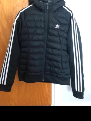 Svart pufferjacka från Adidas - Snygg svart pufferjacka från Adidas med klassiska vita ränder längs ärmarna och Adidas-logga på bröstet. Jackan har huva, dragkedja framtill och två sidofickor. Använd ett fåtal gånger
