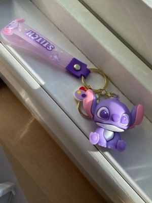 Lila Stitch nyckelring - Söt nyckelring med en lila Stitch-figur och matchande handledsrem i plast där det står 'STITCH' i vitt. Nyckelringen har detaljer i rosa och vitt samt en liten blomma på remmen. Perfekt att hänga på väskan eller nycklarna.