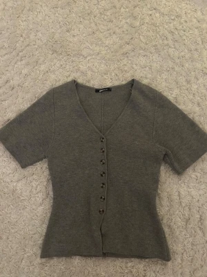 Grå kortärmad kofta Gina Tricot - Stilren grå kofta från Gina Tricot med korta ärmar och v-ringning. Koftan har knappar framtill och är figurnära i modellen. Perfekt att matcha med jeans eller kjol för en enkel och snygg look.