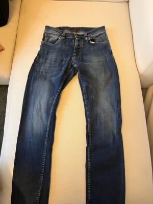 Blåa nudie jeans - Säljer ett par klassiska blå jeans från Nudie Jeans med snygga slitningar och kontrastsömmar. Modellen har fem fickor och en normal passform med raka ben. Materialet är robust denim i bomull och bakfickorna har Nudie Jeans signaturbroderi. I storlek w30/l32