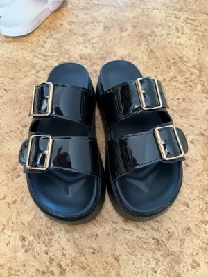 Svarta sandaler med stora spännen - Snygga svarta sandaler med två breda remmar i glansigt material och stora, silvriga metallspännen. Sandalerna har en chunky sula och en modern look som passar till många olika stilar. Perfekta för dig som gillar trendiga och bekväma skor.