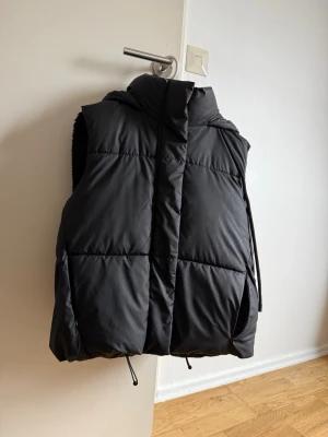 Svart puffer dunväst med huva - Säljer en svart puffer dunväst med huva och dragsko i nederkant. Västen har en oversized look och breda quiltade paneler som ger en modern och trendig stil. Perfekt för lager på lager och enkel att matcha med olika outfits.