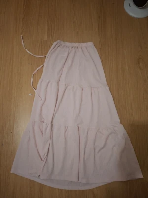 Ljusrosa långkjol med volanger - Söt ljusrosa långkjol med volangdetaljer och knytband i midjan. Kjolen har en avslappnad passform och är perfekt för varma dagar. Tillverkad i mjukt bomullstyg som känns skönt mot huden.