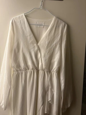 Vit omlottklänning från Sisters Point - Stilren vit omlottklänning från Sisters Point med långa ärmar och v-ringad hals. Klänningen har volangdetaljer framtill och resår i midjan för en snygg passform. Tillverkad i ett lätt, skirt material som ger en luftig känsla. Kostade 499kr ny. 