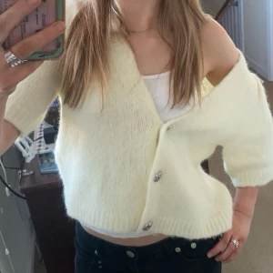 Gul kortärmad stickad kofta - Säljer en ljusgul stickad kofta med korta ärmar och stora silvriga knappar framtill. Koftan har en croppad passform och ribbstickade kanter. Perfekt att slänga över ett linne för en chill look. Så fin, köpt för 700kr