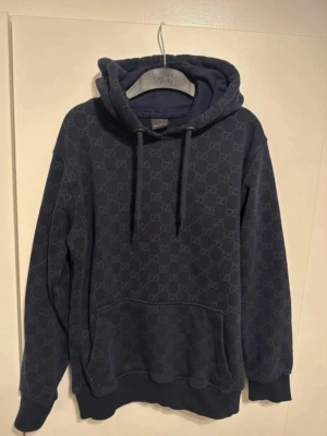 Mörkblå hoodie  - Snygg mörkblå hoodie med diskret GG-mönster över hela tröjan. Klassisk känguruficka framtill, justerbar huva med snören och ribbade muddar. Perfekt för dig som vill ha en stilren och exklusiv look. Materialet känns mjukt och bekvämt.