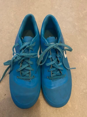 Blå Nike fotbollsskor med dobbar - Säljer ett par blå fotbollsskor från Nike med vita detaljer och klassisk swoosh på sidan. Skorna har snörning och dobbar för bra grepp på planen. Tillverkade i slitstarkt syntetmaterial som tål tuffa matcher. Perfekta för dig som vill sticka ut på fotbollsplanen.