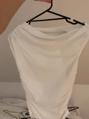 Vit topp med rynkade sidor - Säljer en stilren vit topp med rynkade detaljer längs sidorna och bred, draperad urringning. Modellen är ärmlös och har en tight passform som framhäver figuren. Materialet känns mjukt och stretchigt, perfekt för att skapa en clean look.