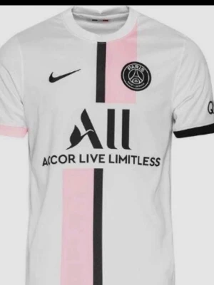 PSG vit matchtröja från Nike - Snygg vit PSG matchtröja från Nike med svarta och rosa vertikala ränder framtill. Tröjan har klubbmärke på bröstet, svart Nike-logga och sponsortryck. Tillverkad i lätt och ventilerande material, perfekt för fotboll eller träning.