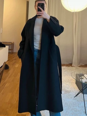 Svart lång kappa med knytskärp - Stilren svart kappa med knytskärp i midjan och klassisk krage. Kappan är lång och har en rak passform, perfekt för att hålla värmen under kyliga dagar. Materialet ser ut att vara ullblandning och kappan har slits bak för extra rörelsefrihet.