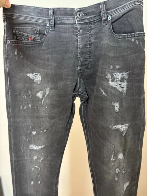 Grå slitna jeans från Diesel - Säljer ett par grå jeans från Diesel D.N.A. med snygga slitningar och lagade detaljer på benen. Jeansen har klassisk femficksmodell, smal passform och cool tvätt. Perfekta för dig som gillar en trendig och avslappnad stil.