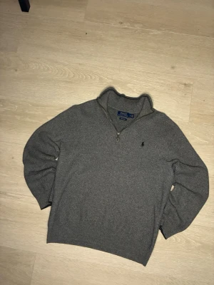 Grå halvzip från Polo Ralph Lauren - Snygg grå stickad tröja från Polo Ralph Lauren med halv dragkedja och klassisk broderad logga på bröstet. Tröjan har hög krage och ribbade muddar vid ärmslut och nederkant. Perfekt till lager på lager och enkel att matcha.