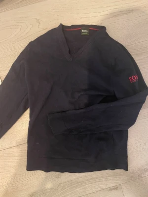 Mörkblå v-ringad tröja från BOSS - Snygg mörkblå v-ringad tröja från BOSS med diskret logga broderad i rött på ärmen. Tröjan har långa ärmar och klassisk passform, perfekt för en stilren look. Materialet känns mjukt och bekvämt. Använd 5 gånger.