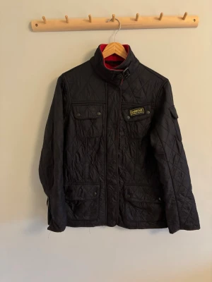 Svart quiltad jacka från Barbour - Snygg svart quiltad jacka från Barbour med hög krage och spänne. Jackan har flera fickor med knappar och Barbour-logga på bröstet. Perfekt för dig som vill ha en klassisk och stilren look med lite edge.