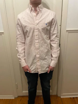 Randig skjorta från morris - Säljer en vit och ljusrosa randig skjorta med lång ärm och blå broderad detalj på bröstet. Skjortan har klassisk krage och knäppning framtill. Perfekt för dig som gillar stilrena och fräscha plagg.