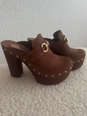  Nya. Bruna platåclogs med gulddetaljer - Snygga bruna clogs med hög platåklack från Zara. Skorna är i skinn med guldfärgade nitar runt sulan och dekorativ gulddetalj framtill. Perfekta för dig som vill sticka ut med en retrovibe.