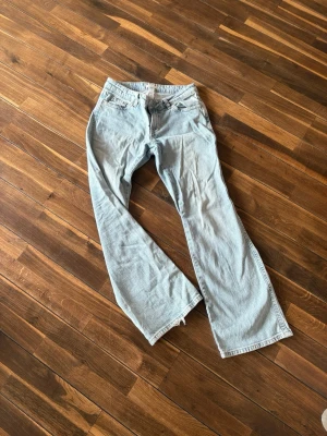 Ljusblå bootcut jeans - Säljer ett par ljusblå bootcut jeans med klassisk femficksdesign och rå kant längst ner på benen. Jeansen har hög midja och är tillverkade i denim med en avslappnad passform. Perfekta för en trendig och avslappnad stil.