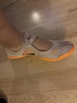 Vibram FiveFingers beige tåskor - Säljer ett par Vibram FiveFingers tåskor i beige med orange sula och detaljer. Skorna har individuella tår, kardborreband över foten och är tillverkade i ett flexibelt textilmaterial. Perfekta för barfotakänsla vid träning eller promenader.