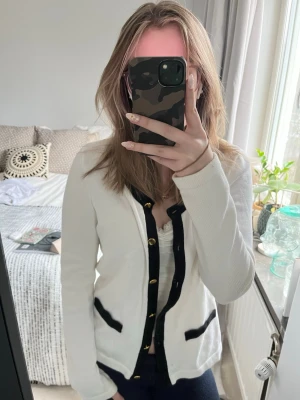 Vit kofta med svarta detaljer H&M - Stilren vit kofta från H&M med svarta kontrastkanter och guldfärgade knappar. Koftan har två dekorativa fickor framtill och lång ärm. Perfekt för en chic och enkel look. I storlek S och från H&M