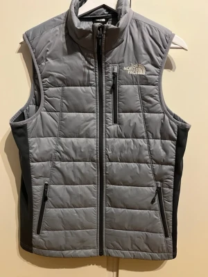 Grå dunväst från The North Face - Snygg grå dunväst från The North Face med hög krage och dragkedja framtill. Västen har tre fickor med dragkedja och diskret logga på bröstet och ryggen.                Pris kan diskuteras                                                                          Quiltad design och svarta sidopaneler ger en sportig look. Perfekt för lager-på-lager under kyliga dagar.