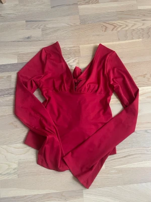 Röd omlottopp med knyt och v-ringning - Trendig röd omlottopp med lång ärm och djup v-ringning. Toppen har ett snyggt knyt baktill som ger en unik look. Perfekt för dig som vill sticka ut lite extra. Tillverkad i mjukt och stretchigt material för skön passform.