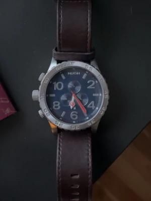 Nixon klocka med brunt läderband - Stilren klocka från Nixon med mörkblå urtavla, silverfärgat boett och tydliga siffror. Klockan har tre mindre urtavlor och en röd sekundvisare. Armbandet är i mörkbrunt läder och ger en klassisk look. Den är äkta, köpt för 4000 men har knappt använt den, den har bara legat i en klock burk,