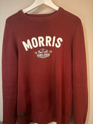 Vinröd stickad tröja från Morris - Snygg vinröd stickad tröja från Morris med vit broderad logga och brittisk flagga på bröstet. Tröjan har rund halsringning, långa ärmar och ribbade muddar. Perfekt för dig som gillar klassisk och stilren design.