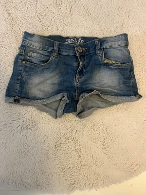 Blå jeansshorts från Lyle - Säljer ett par klassiska blå jeansshorts från Lyle med upprullade benslut och fem fickor. Shortsen har normal passform och är tillverkade i bomull med en snygg tvättad look. Perfekta för varma dagar!