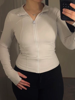 Vit figurnära ziptröja - Snygg vit ziptröja med figurnära passform och långa ärmar. Tröjan har dragkedja framtill och en liten krage. Perfekt för en sportig och clean look.