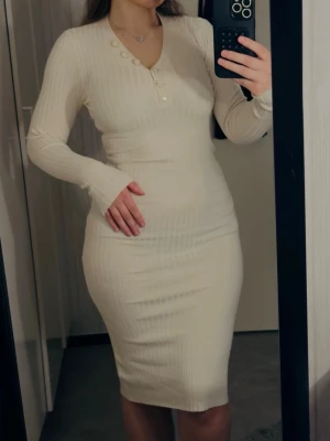 Ljus ribbad långklänning med knappar - Snygg ljus ribbad långklänning med lång ärm och v-ringning med dekorativa knappar framtill. Klänningen har en kroppsnära passform och är tillverkad i ett mjukt, stretchigt material som sitter bekvämt. Perfekt för dig som gillar stilrena och enkla plagg.