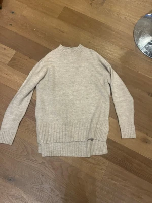 Beige stickad tröja med slits - Mysig beige stickad tröja med lång ärm och ribbade muddar. Tröjan har en liten slits i sidan och en högre halsringning. Perfekt för lager på lager och enkel att matcha till olika outfits.
