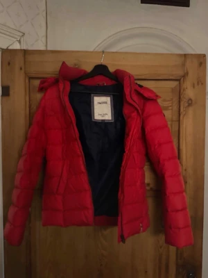 Röd pufferjacka från Tommy Hilfiger  - Snygg röd pufferjacka från Tommy Hilfiger med huva och svart innerfoder. Jackan har quiltad design, dragkedja framtill och två sidofickor. Perfekt för kalla dagar och ger en cool streetstil. Materialet är syntetiskt och jackan är vadderad för extra värme.